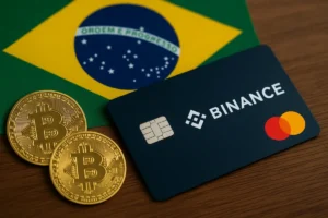 Cartão cripto da Binance com bandeira Mastercard ao lado de duas moedas de Bitcoin, sobre mesa, com a bandeira do Brasil ao fundo.